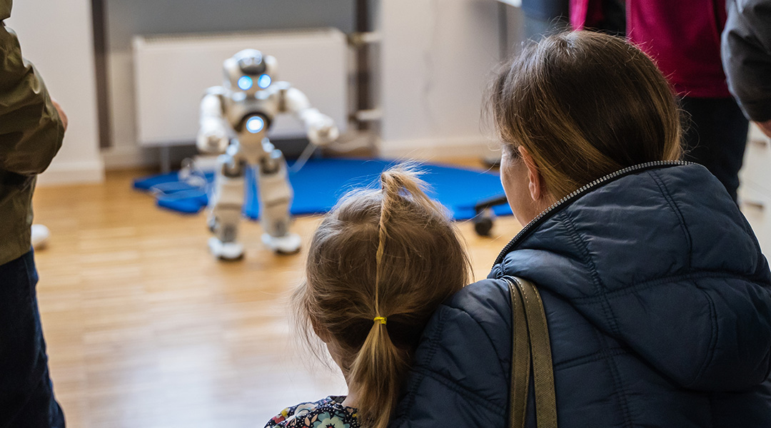 Eine Mutter schaut mit ihrer Tochter zusammen im BayernLab Forchheim dem Roboter NAO zu, wie er eine Fitness Übung präsentiert. 