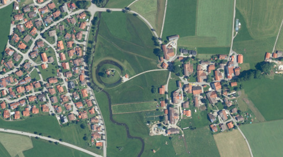 Das Farbluftbild zeigt Wiesen, Ortschaften, einen Bach  und in der Mitte einen kreisrunden Hügel mit einem Haus.